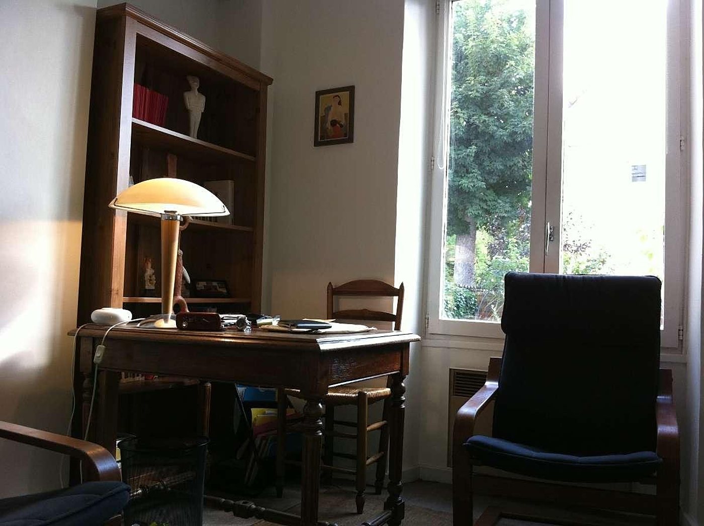 Pièce lumineuse meublée d'un bureau en bois avec une lampe champignon allumée, d'une bibliothèque, d'un fauteuil bleu foncé et d'une grande fenêtre donnant sur un arbre.