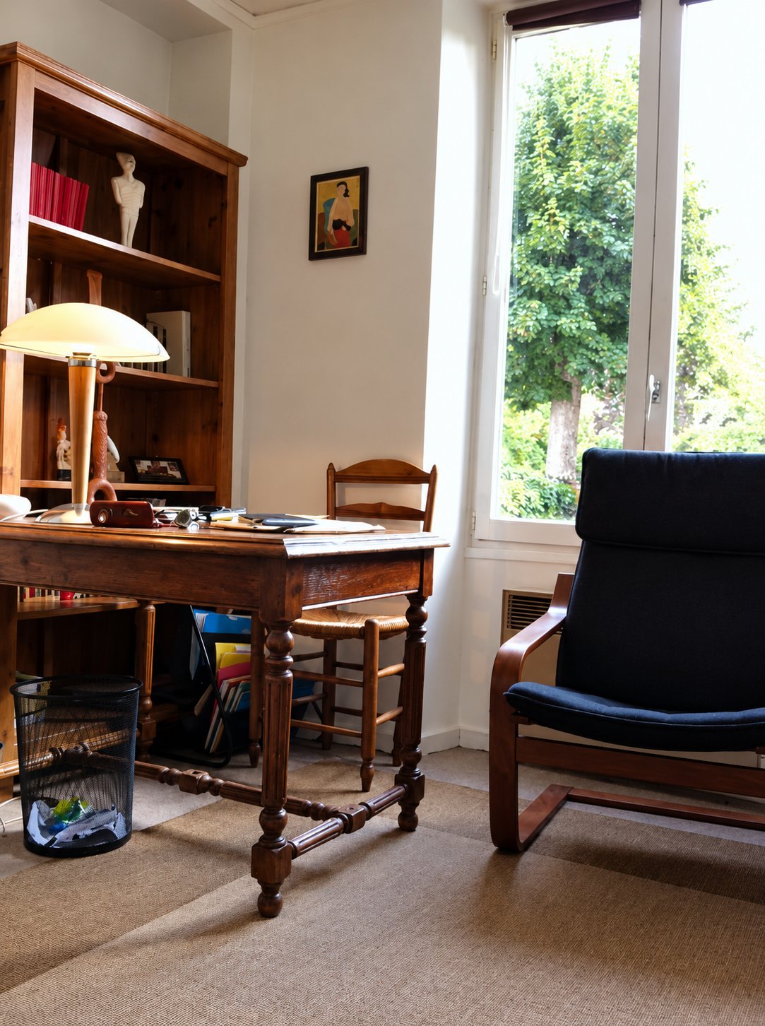 Un bureau ancien en bois avec une lampe, une chaise paillée et un fauteuil bleu devant une grande fenêtre donnant sur un arbre, à côté d'une bibliothèque.