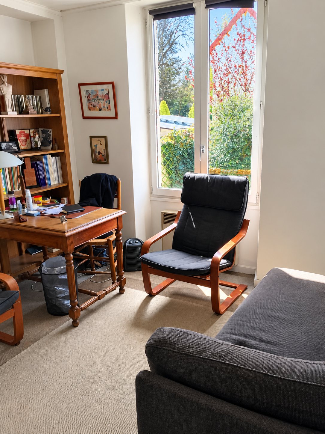 Pièce claire aménagée en cabinet avec un bureau en bois, une bibliothèque, un fauteuil noir près de la fenêtre et un canapé gris au premier plan.