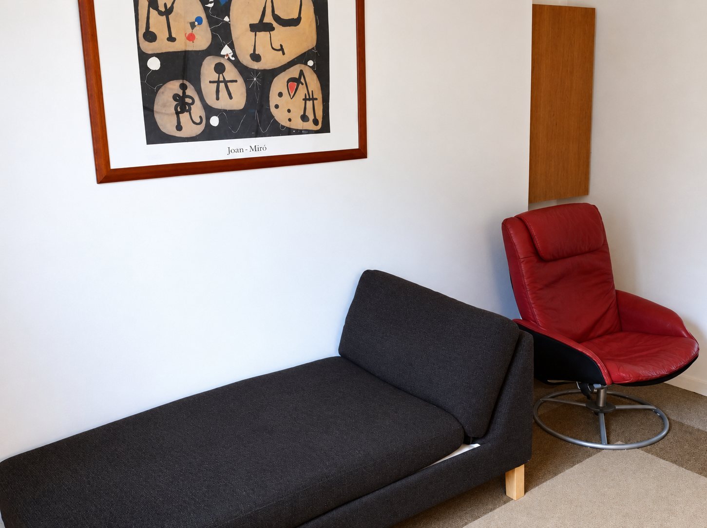 Une méridienne grise et un fauteuil pivotant rouge sont installés dans un coin de pièce, sous une affiche encadrée de Joan Miró.