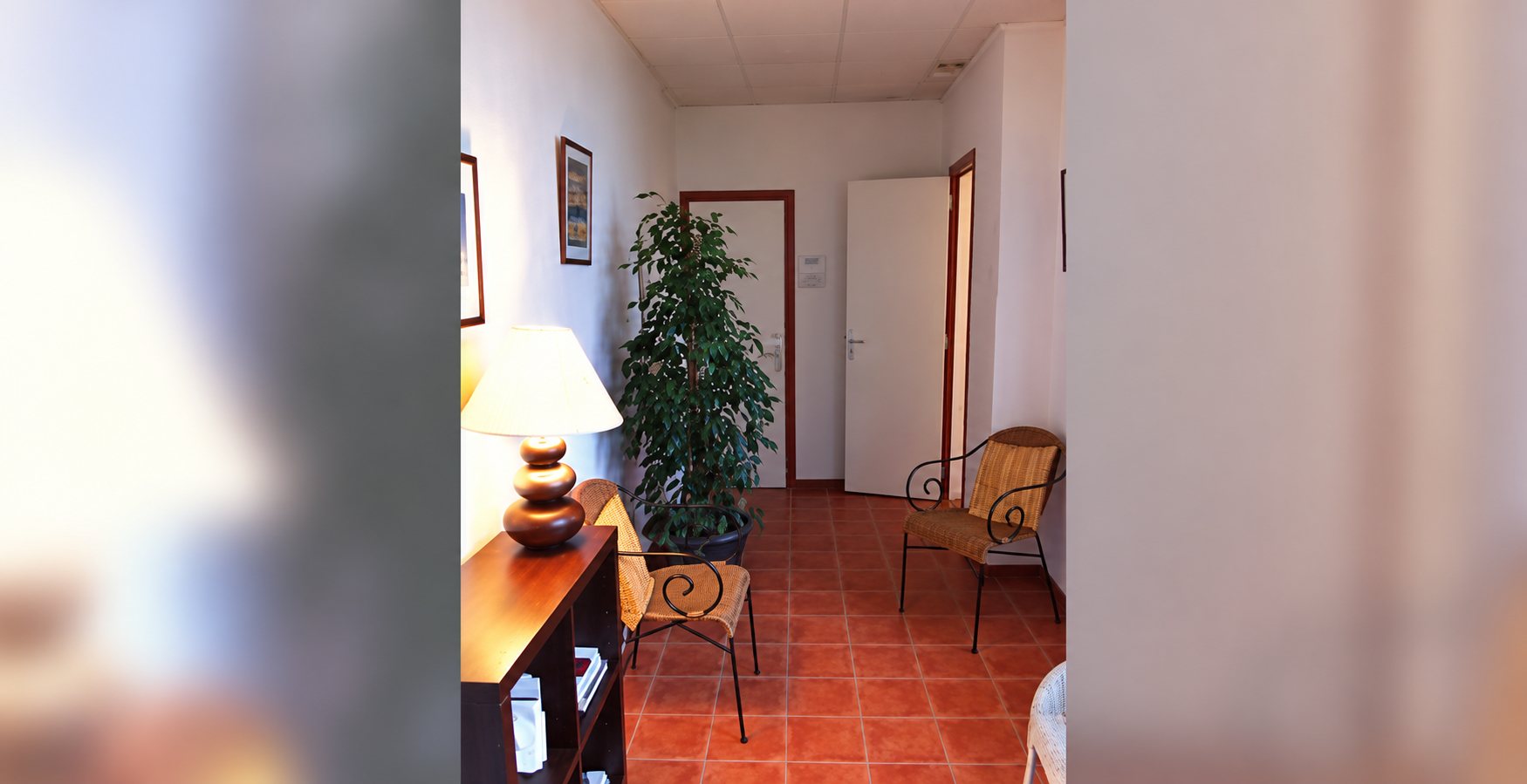 Un couloir aux murs clairs et au sol en carrelage rouge, avec une grande plante verte, une lampe posée sur un meuble en bois et des fauteuils en fer forgé.