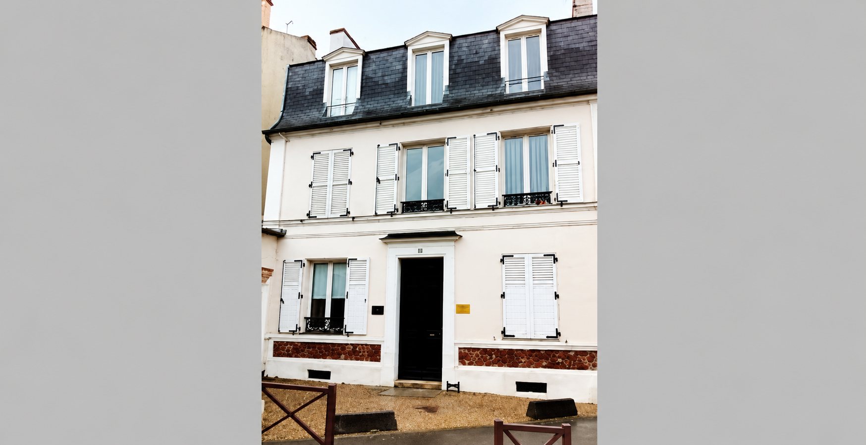 Façade d'une maison de ville à trois niveaux aux murs clairs, volets blancs, lucarnes et toit en ardoise, avec une porte sombre au centre.