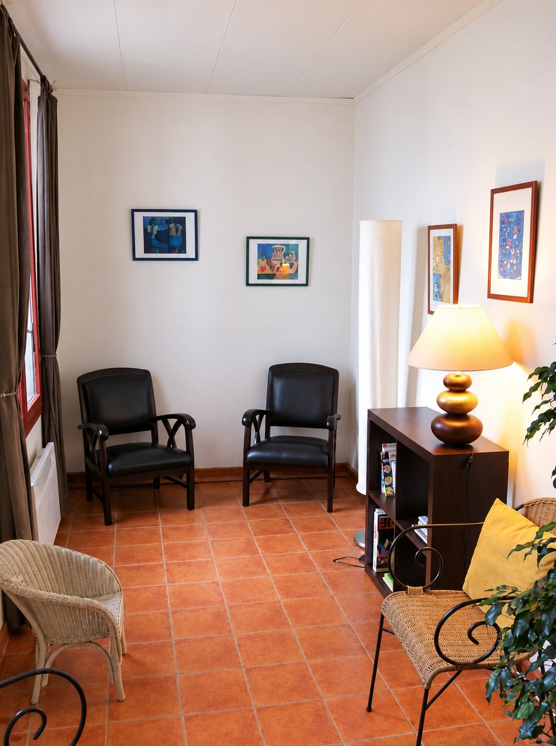 Une salle d'attente aux murs clairs avec deux fauteuils en cuir noir, une lampe allumée sur une étagère, des tableaux et un sol en carrelage orangé.